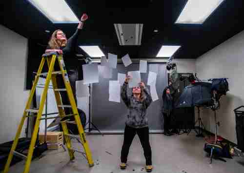 Three people hang paper from a photo studio ceiling. One stands on a ladder on the left, another in the middle. A third person adjusts a light at the right.