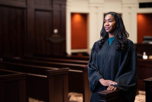 Kyra stands in the court room wearing a black robe and looking off to the left