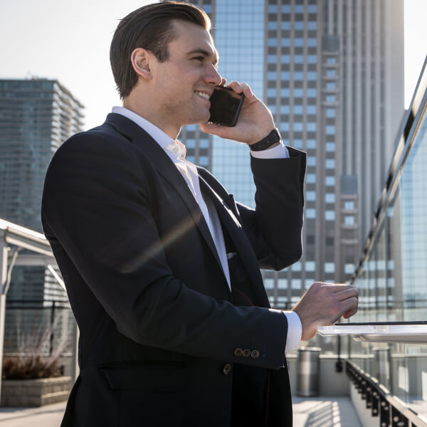 Alumnus Jason Growhowski stands in front of a wall-mounted desk unit while wearing a suit in a big city holding a phone.