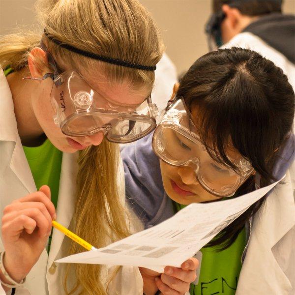 Two people wearing goggles peer at a piece of paper that one person is holding. That person is also holding a pencil.