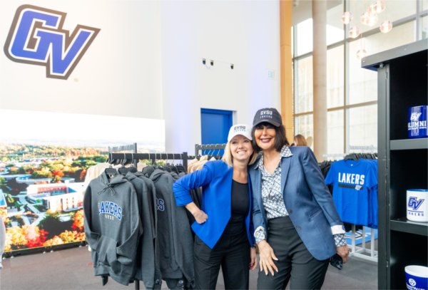 Two women in professional attire, pose together wearing baseball caps inside a store. 