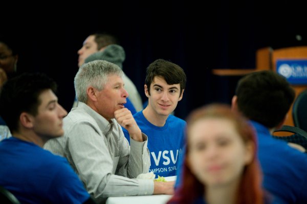 student in blue shirt listens to man in gray sweater talk at table