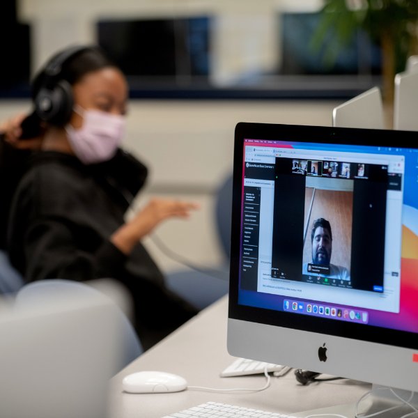 computer in forefront with image of student, Grand Valley student in background with headphones on and a pink face mask