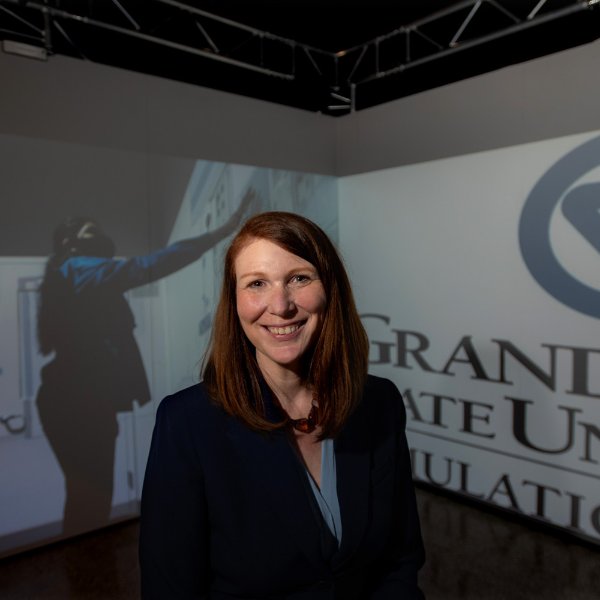 Katie Branch seated in front of immersive technology in background showing a projection of a student touching a wall and the Grand Valley State University, simulation center logo on the other wall