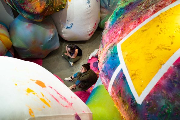 Two people sit on the floor of an art gallery surrounded by an installation of giant inflated colorful balls of painted fabric. 