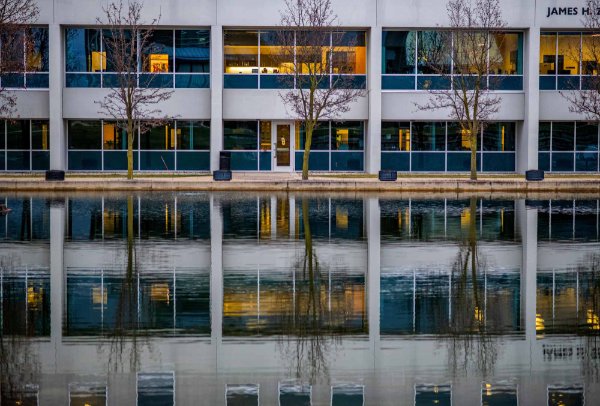 Geometric rectangular spaces of a building shine blue and orange and reflect on the surface of a pond. 