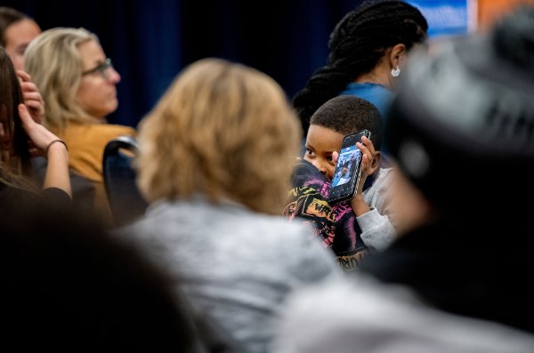 A young child holds up a phone to show the people behind him. He is seen through others in a crowded room.  