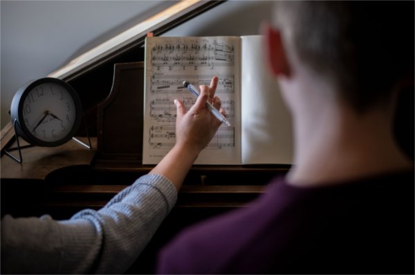 The back of a student is seen and the arm of a piano teacher, pen in hand, gesturing toward a music book.