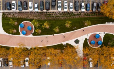 View from drone of curving sidewalk with people walking on it and cars parked in rows.