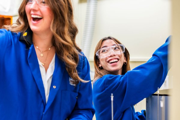 Two people dressed in blue lab coats and safety glasses laugh and smile in a lab setting. 