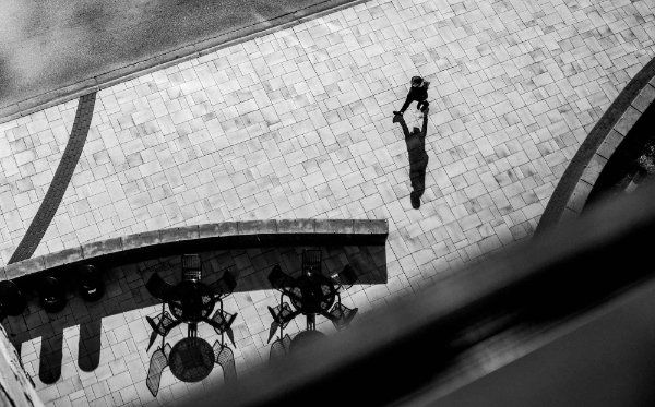  Viewed from above from a window, a person walks along a patterned sidewalk, their shadow prominently showing on the ground. 
