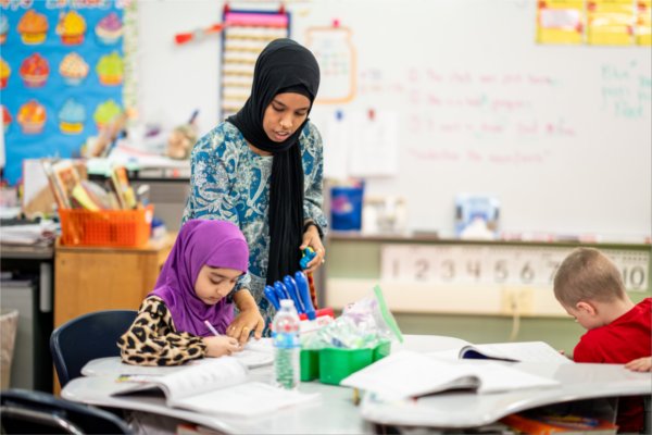 teacher in a hajib stands over the desk of a young student, also in a hajib