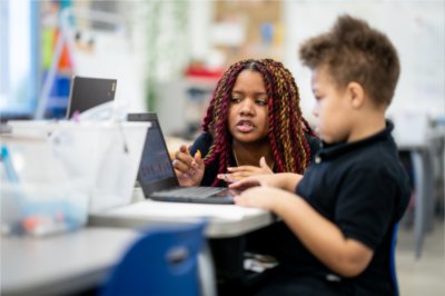 teacher kneels by desk of second grader who is looking at a laptop