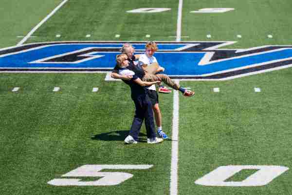 Two people carry their father on the 50-yard line of a football field. 