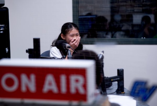 A person covers their mouth with their hand while laughing. A sign with the words "On Air" is in the foreground.