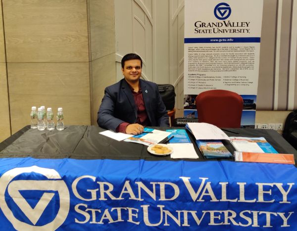 Kirthi Kondapalli sits behind a table during a recruitment event in India. Banner behind him and table cloth reads Grand Valley State University.