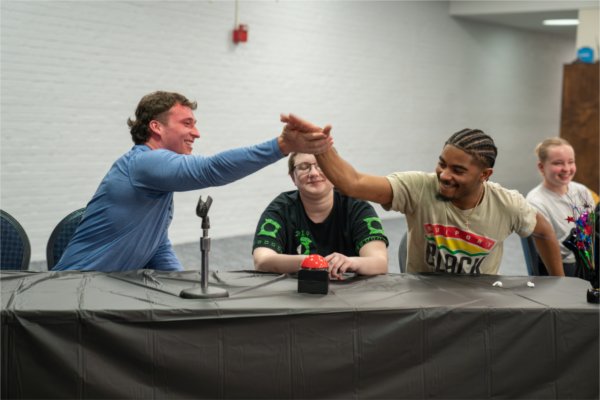 students share a high five at a table while student in middle smiles during a quiz bowl