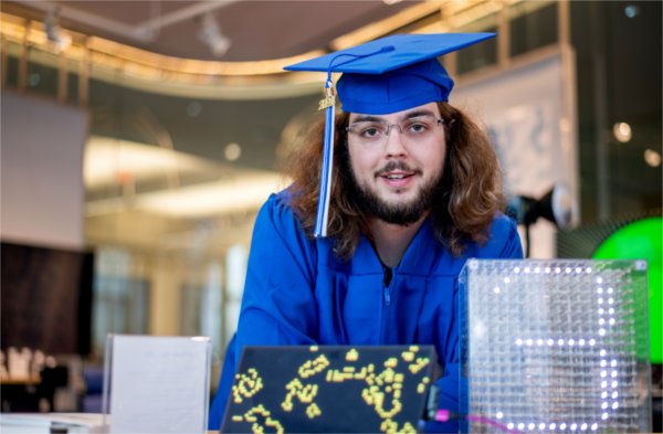A person wearing a cap and gown smiles while looking at the camera. Some computing-related devices are in front of the person.