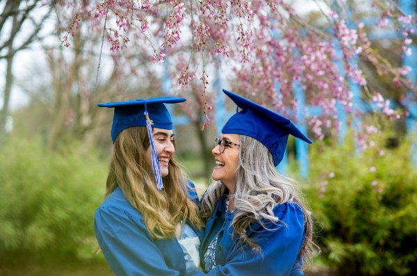 Two people wearing caps and gowns smile while looking at each other. The pink blossoms of a tree hang overhead.