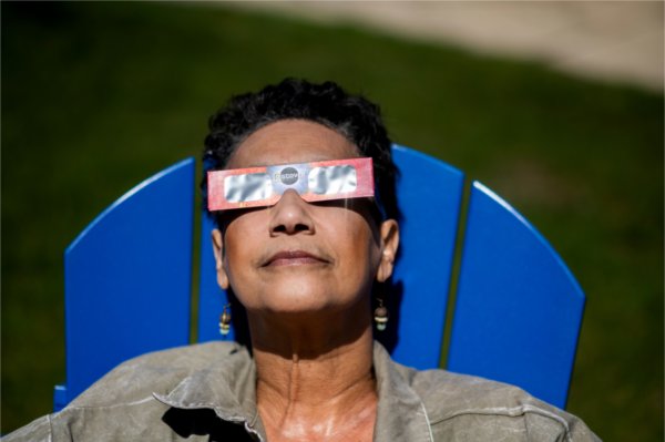 A person wearing special eclipse glasses leans their head against the back of a blue chair while looking skyward.