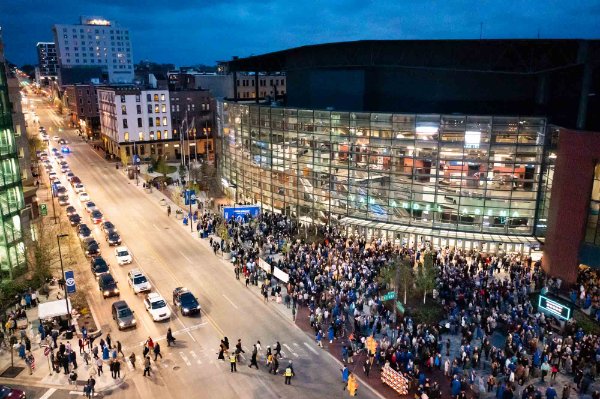  Photo from above showing a street with cars, a lot of people in caps and gowns coming out of a lit up arena. 