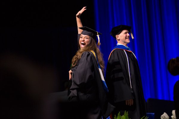 A person in a cap and gown celebrates on stage with one hand high in the air and the other clutching their diploma.