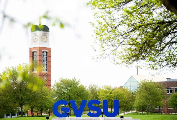 Big blue letters that say "GVSU" stand out amongst a campus of green trees, with buildings and a clock tower in the background. 