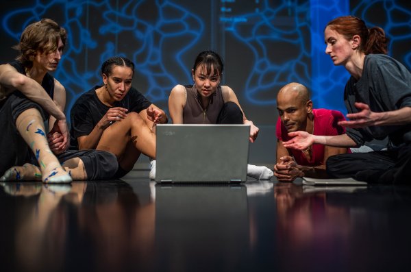  Five Grand Rapids Ballet dancers sit on the stage watching something on a laptop that is sitting on the floor in front of them. 