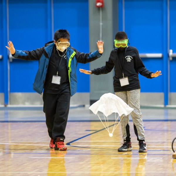 Two people wearing goggles hold their hands in the air as they watch a small parachute lift off from a gymnasium floor.