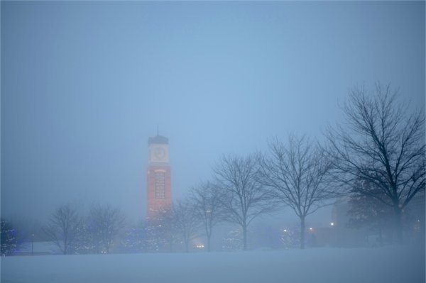  A carillon tower is seen past a row of trees in a foggy scene on a college campus at dawn. 