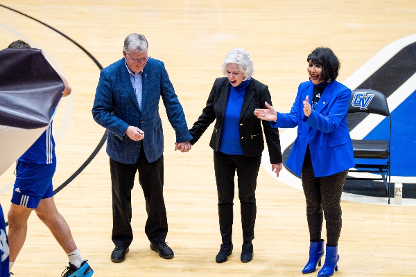  A university president wearing blue claps as a basketball court named after a former university president, pictured holding hands with his wife, is unveiled. 