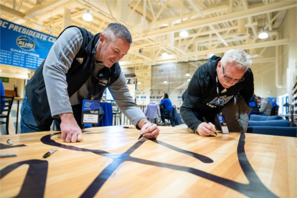 GVSU head football coach Scott Wooster, left, signs a banner congratulating President Emeritus Thomas J. Haas for the Fieldhouse Arena basketball court being named in his honor. 