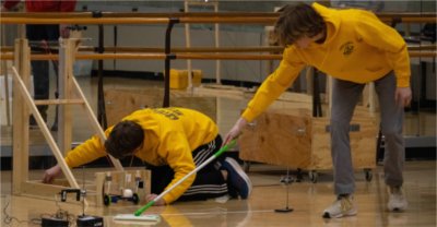 two students in yellow work on floor during robotics event, one holding swifter mop