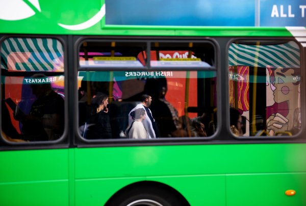 The windows of a green bus reflect people in the city and signs, reading backward, "Irish burgers" and "breakfast."