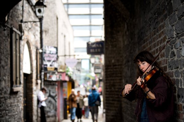 A person plays the violin while leading against a brick wall with a city sidewalk in the background.