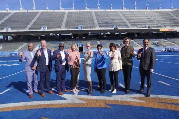 Nine people stand in a line in a stadium with a blue football field. They are using their hands to signal the number "four."