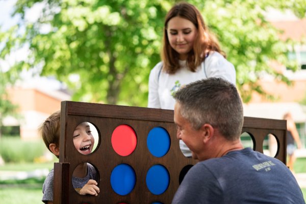  Child sticks his smiling face through the hole of a giant connect four set while his sister and father laugh and smile at him. 