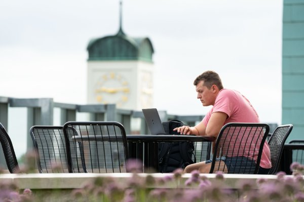 A person sits on a rooftop terrace with flowers in the foreground and the top of the clock tower in the background.