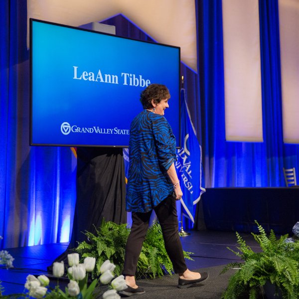 LeaAnn Tibbe walks across a commencement stage, the screen behind her reads her name: LeaAnn Tibbe and the Grand Valley State University wordmark