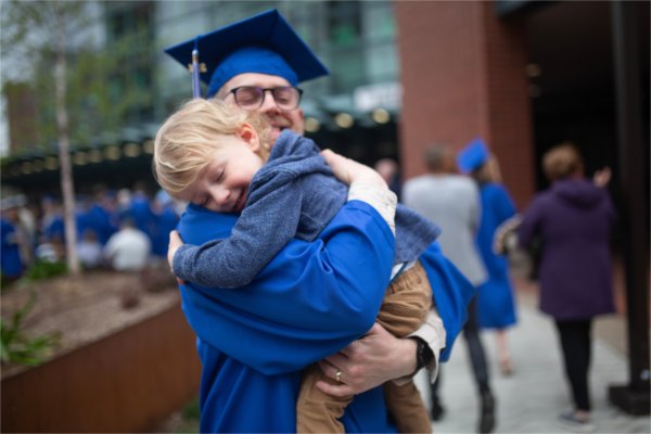  A graduate wearing cap and gown holds his toddler son before Commencement.