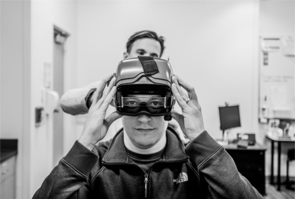 Justen Knape, a graduate student, dons an immersive headset in the Community Hearing Clinic, in Finkelstein Hall, with the help of faculty member Colton Clayton, who is standing behind him.