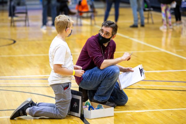 An adult wearing a mask speaks to a child wearing a mask. Both are kneeling on a gymnasium floor; the adult is holding a clipboard.