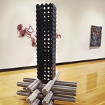 Black wooden pillar with circles cut out on top of stacked cement rails in gallery space