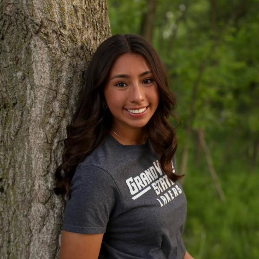 Lizette leaning against a tree wearing a grey t-shirt that says Grand Valley State Lakers on it