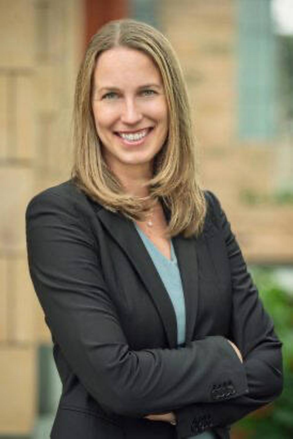 A blonde woman in a blazer smiling at the camera with crossed arms.