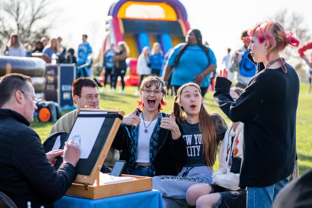 A group of students sitting outside and posing for a caricature artist. There's a bouncy house and lots of other students in the background.