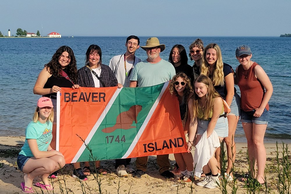 Students and professor standing on the Beaver Island shore, holding a flag, with Lake Michigan in the background