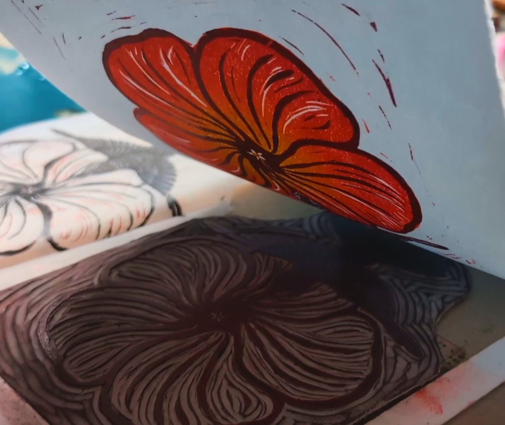 Linoleum block with a flower carved into it. A print of the flower, which is orange, is being pulled up.