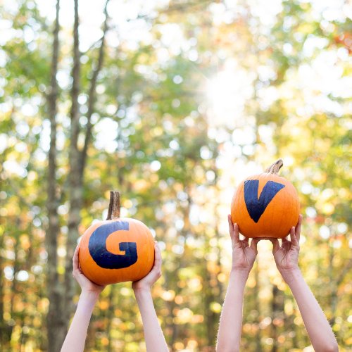 Two pumpkins being lifted into the air, one with a hand painted "G" and the other with a hand painted "V"