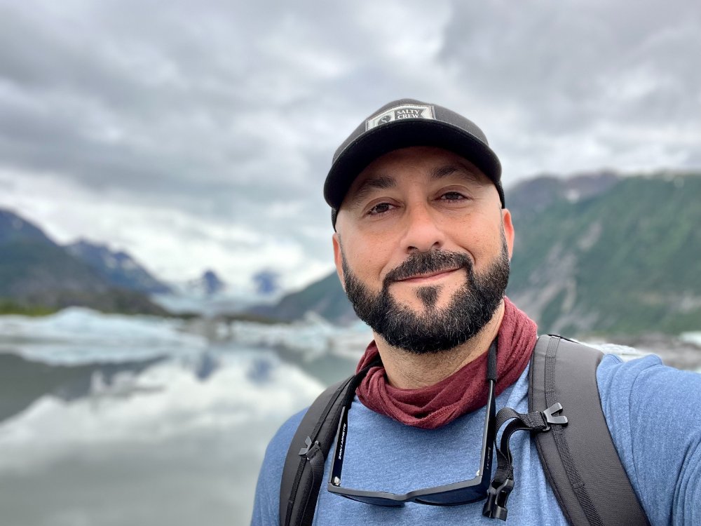 a bearded man in a baseball cap stands in the mountains